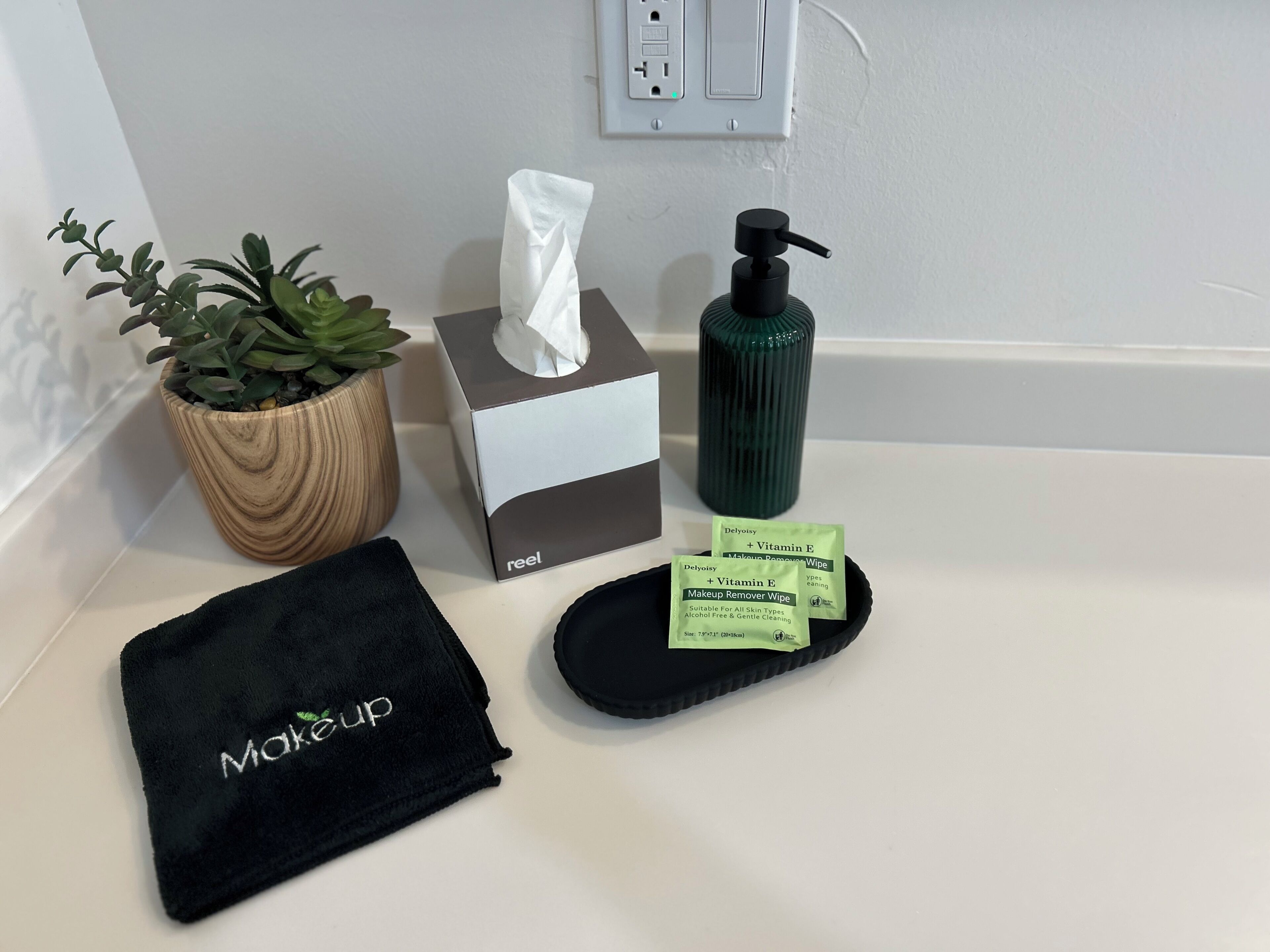 Un meuble-lavabo dans une salle de bain, avec une boîte de mouchoirs, un lingette démaquillante, une plante en pot, une serviette noire et une bouteille verte.