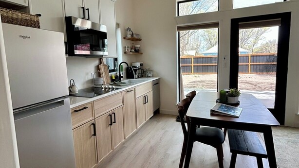 Una cocina moderna con refrigerador, microondas y alacenas de madera. Hay una mesa de comedor con sillas y vistas al exterior a través de amplias ventanas.