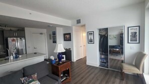 Ein modernes Wohnzimmer mit einem grauen Sofa, einem kleinen Holztisch, einer Lampe und einem Spiegel. Es gibt eine Küchenzeile mit einem Kühlschrank und einer Spüle.