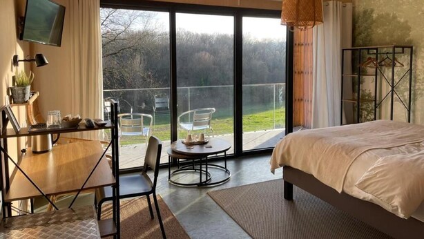 A bedroom with a bed, a desk, a chair, a TV, and a balcony with a view of trees. - Les libellules (Gémil)