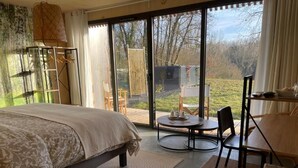 A bedroom with a bed, a round table, chairs, a small table with items, and a view through large windows. - Les libellules (Gémil)