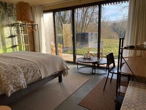 A bedroom with a bed, a round table, chairs, a small table with items, and a view through large windows. - Les libellules (Gémil)