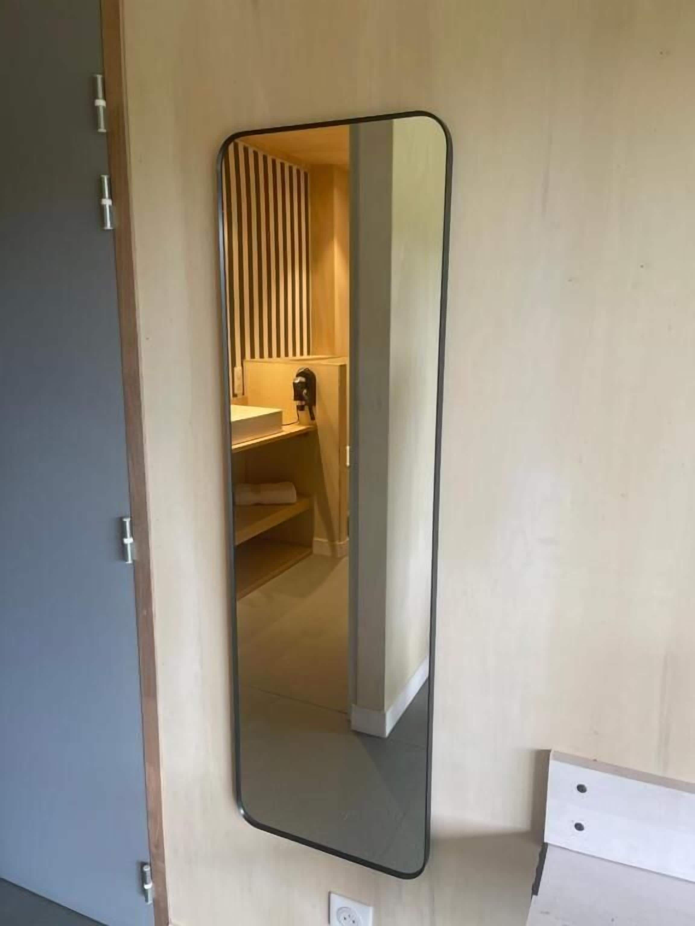 Un espejo sujeto a la pared con un panel de madera, un enchufe y una vista parcial de un baño con un perchero.
