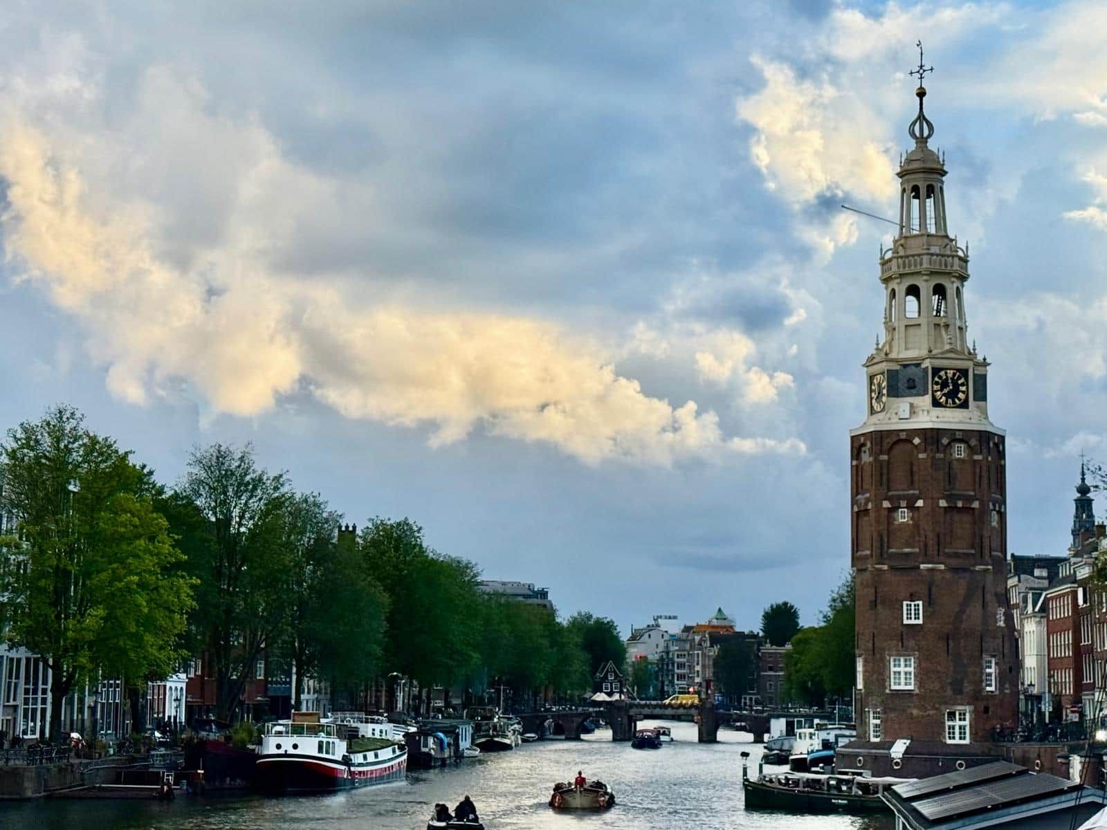 A historic tower with a clock, a river with boats, and a bridge in a cityscape.