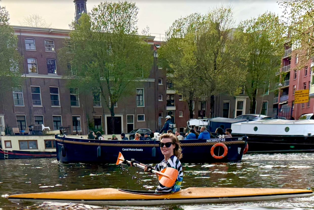 A person in a kayak on a canal with buildings and boats in the background.