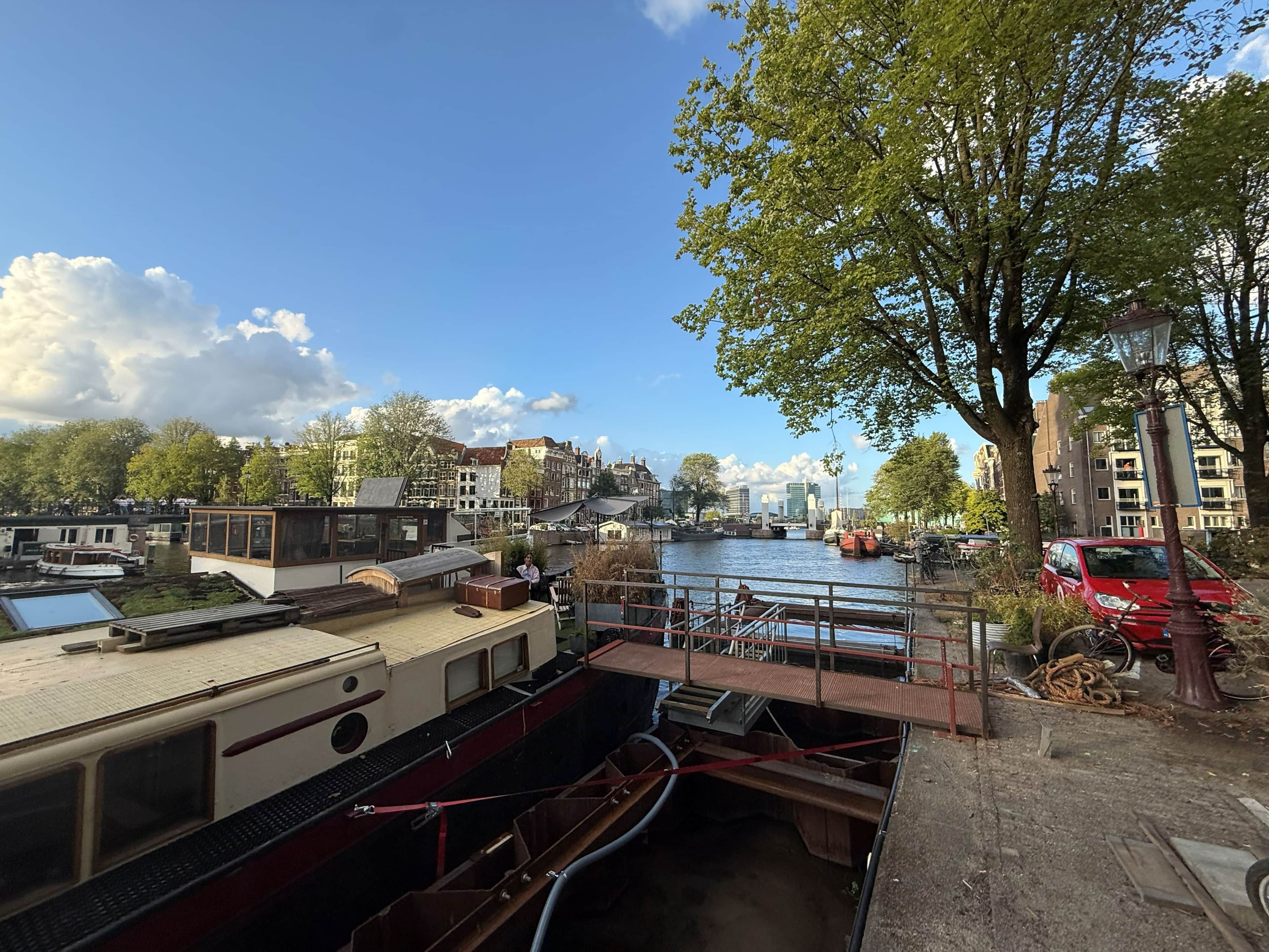 Blick auf einen Fluss mit vertäuten Booten, einer Fußgängerbrücke und Gebäuden im Hintergrund.