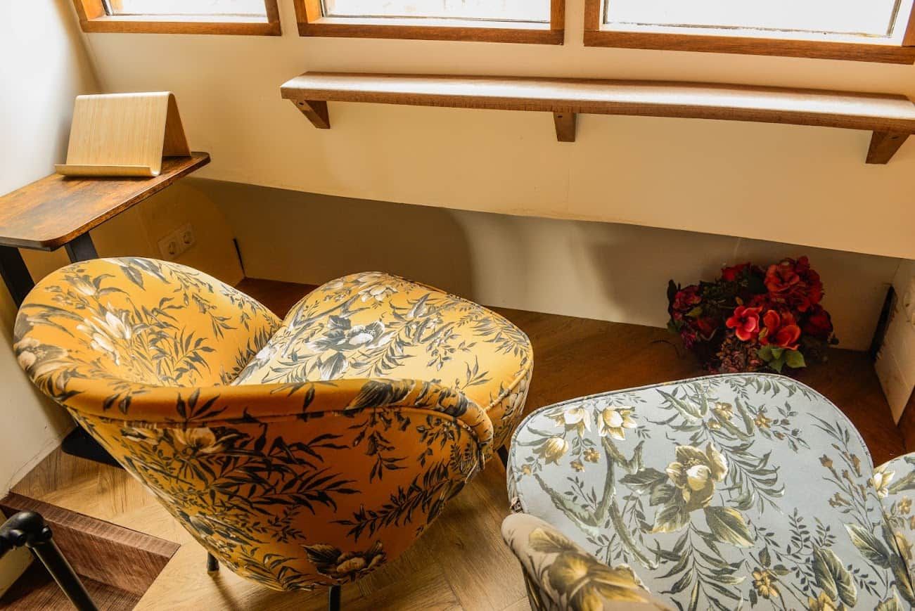 A corner of a room with two upholstered chairs with floral patterns, a small table with a book, and a vase with red flowers.