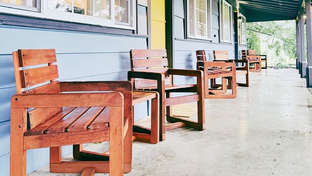 Un espace extérieur abrité, doté de bancs et de tables en bois, d’un sol en béton et d’un bâtiment avec de grandes fenêtres et une porte jaune.
