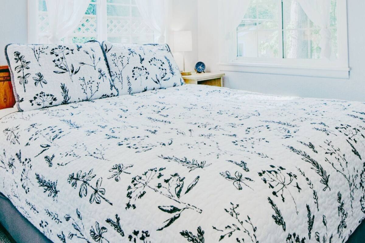 A bedroom with a bed, two windows with sheer curtains, a bedside table with a lamp, and a view of greenery outside.
