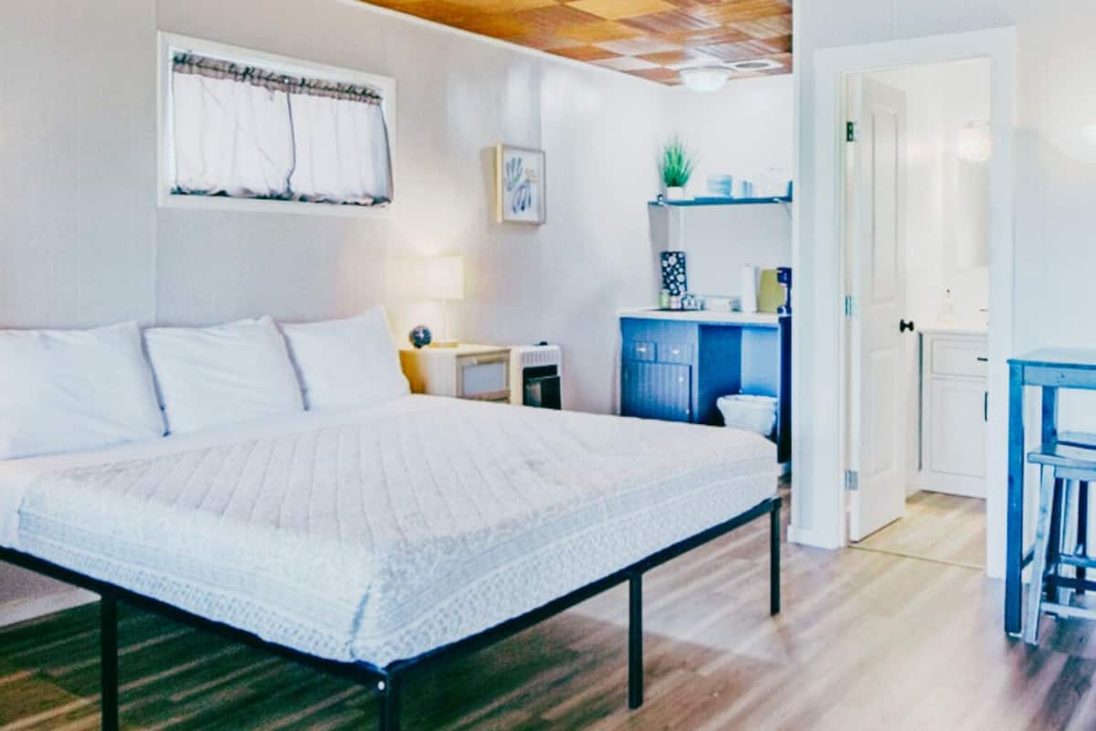 A bedroom with a large bed, wooden flooring, a checkered ceiling, a small kitchenette, and a bathroom visible through an open door.