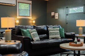 A dark-themed living room with a leather sofa, two side tables with lamps, a small round table with a candle, and a window with blinds.