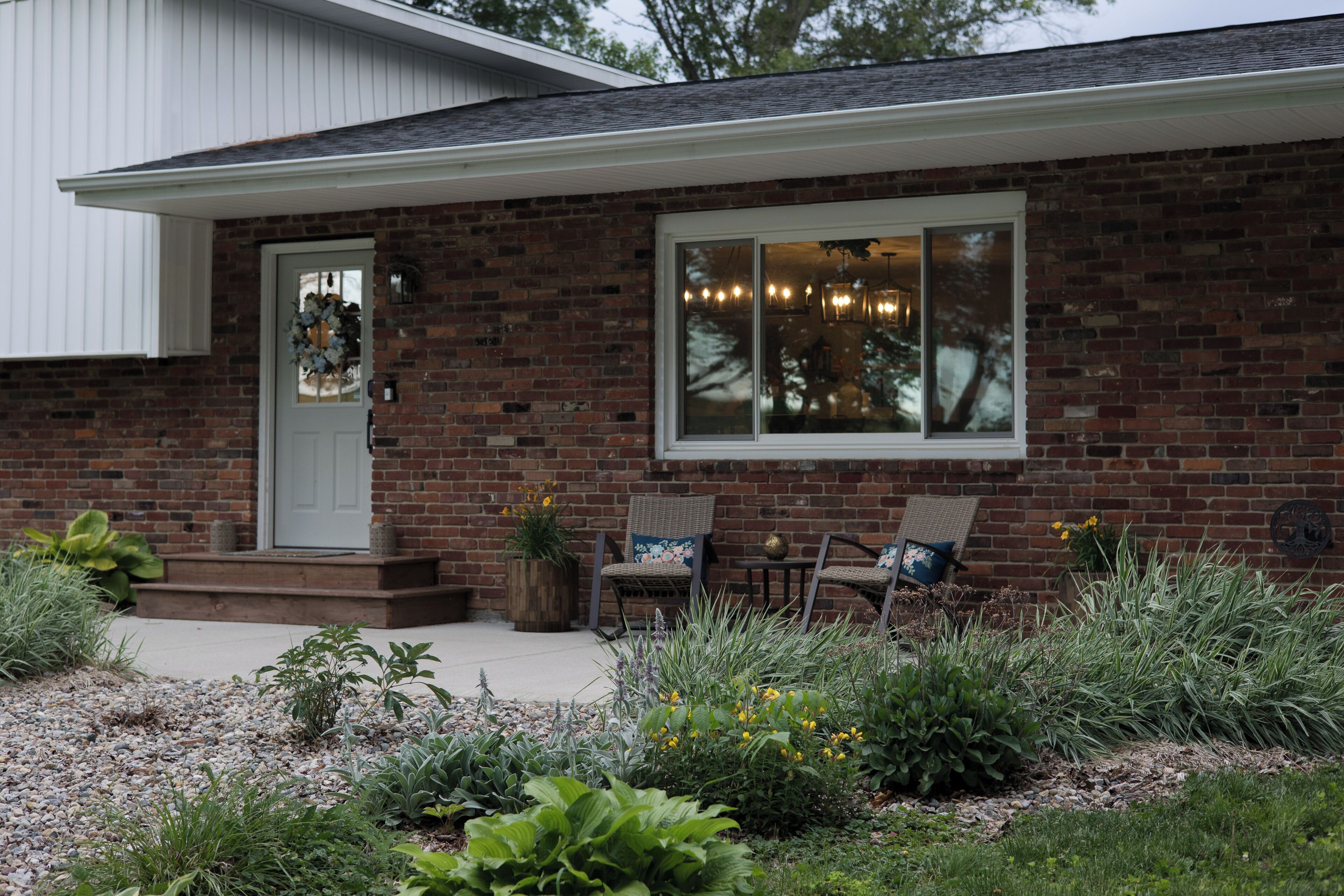 A brick house with a front porch, a door with a wreath, two wicker chairs, and a window with lights.