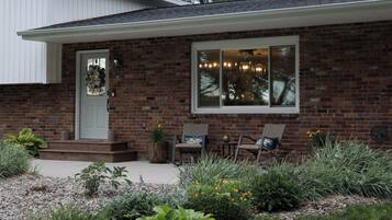 A brick house with a front porch, a door with a wreath, two wicker chairs, and a window with lights.