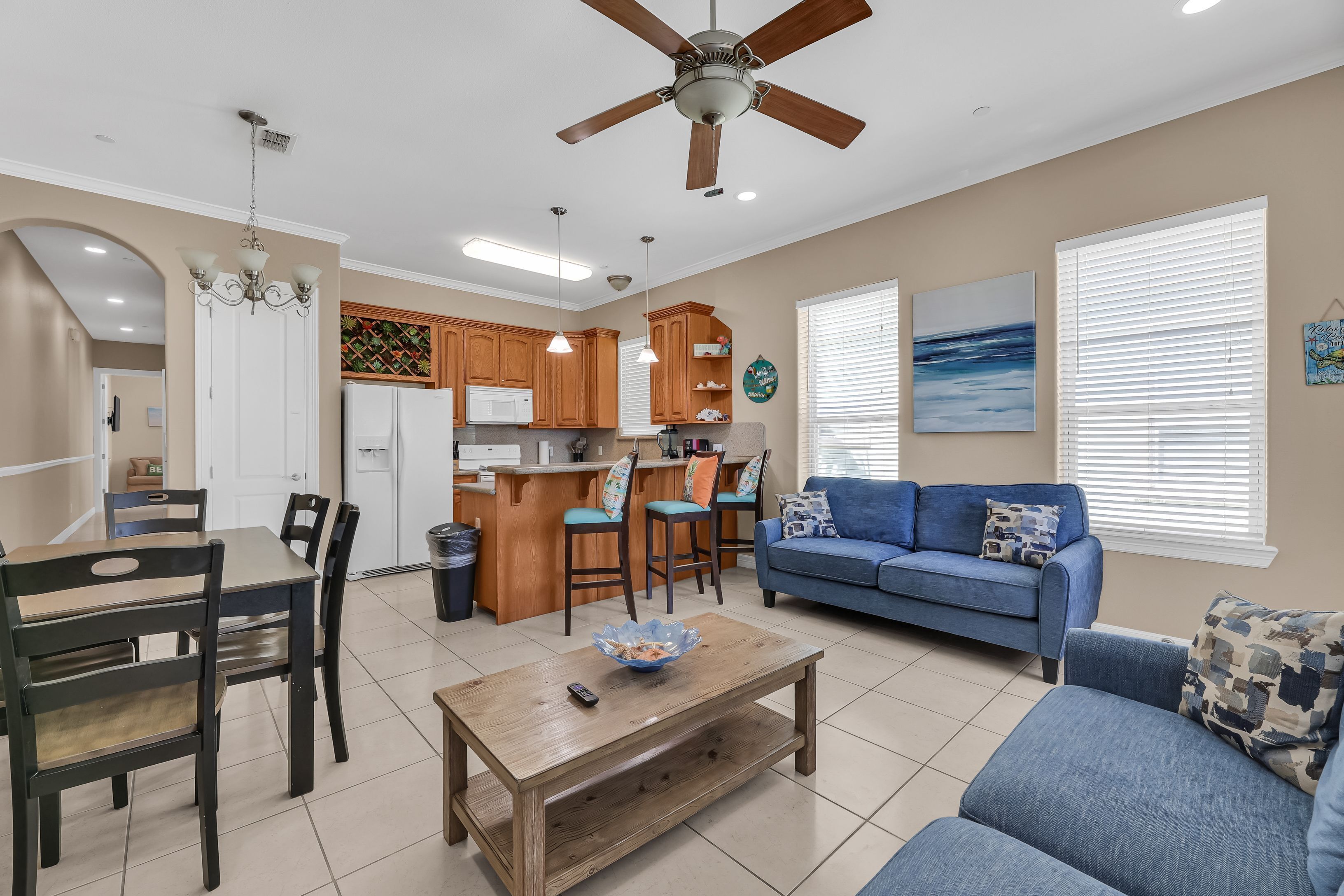 A living room with a blue sofa, wooden coffee table, and a kitchen area with wooden cabinets and a white refrigerator. - Beachfront Condo with Private Balcony Pool (South Padre Island)
