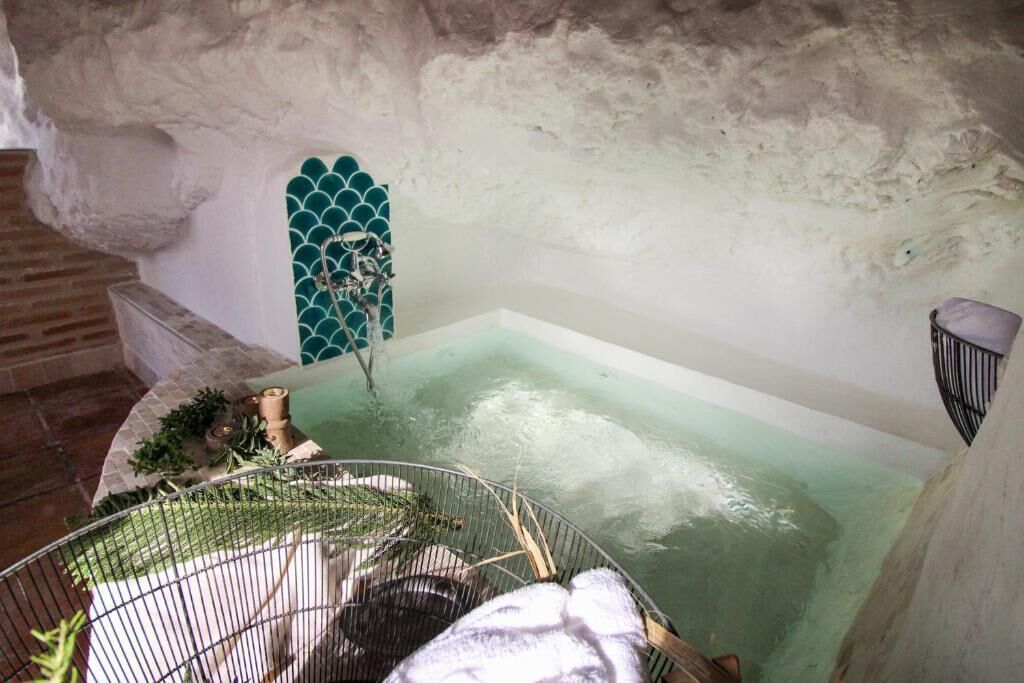 Une baignoire dont l’eau a une teinte verdâtre, une cage en métal avec une serviette, et un mur de pierre rustique.