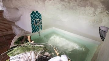 Une baignoire dont l’eau a une teinte verdâtre, une cage en métal avec une serviette, et un mur de pierre rustique.