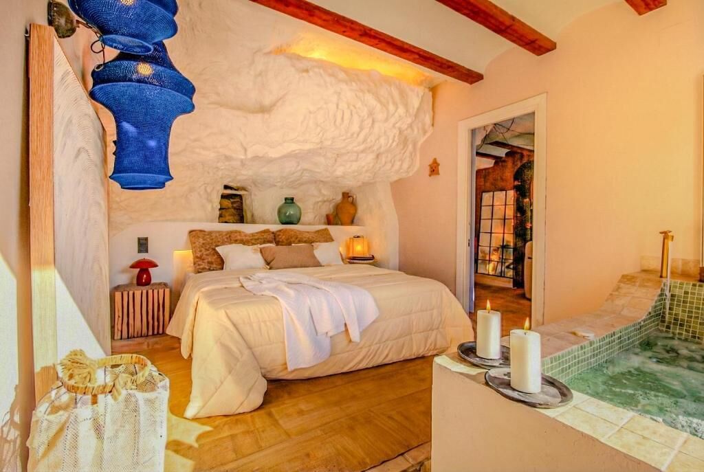 Un dormitorio con cama, mesita de noche, lámpara, estante con adornos y vista a una pared de piedra y una ventana.