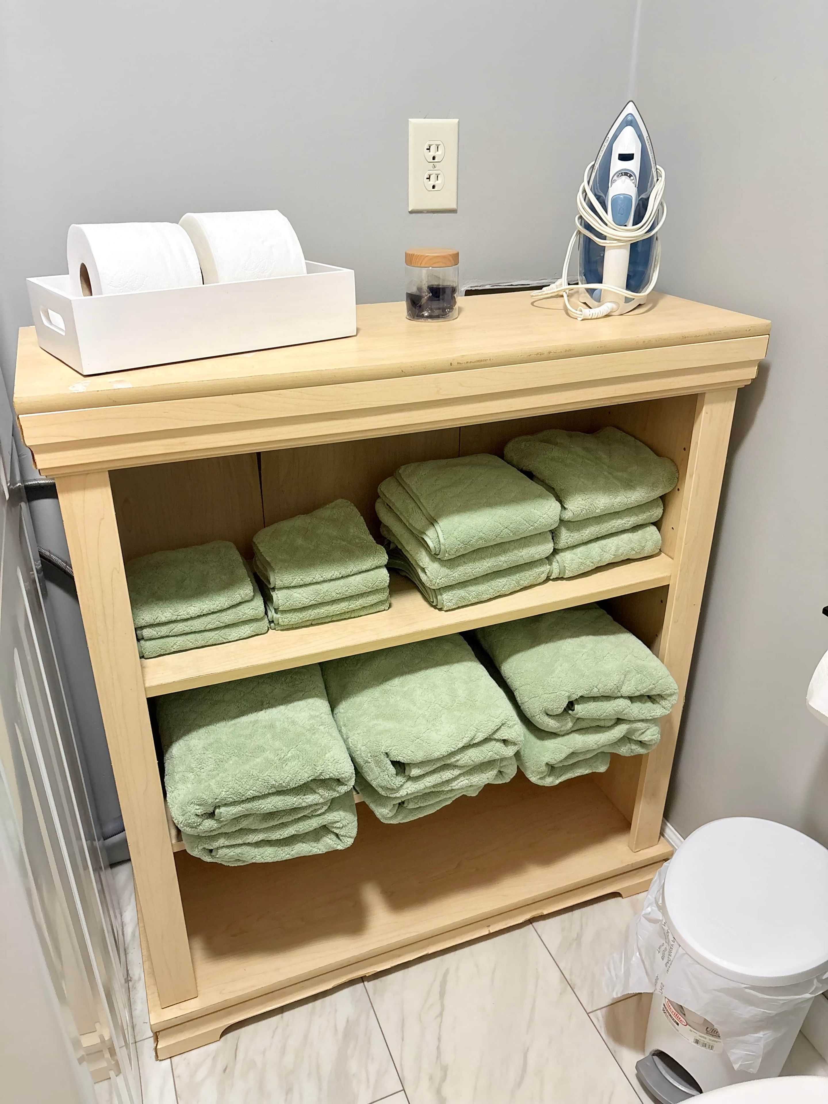 A wooden shelf with neatly folded green towels and a white box of toilet paper.
