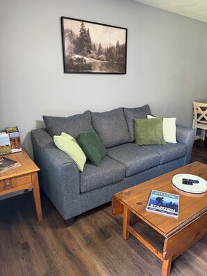 A grey sofa with green and white cushions, a framed landscape painting, a wooden coffee table with a welcome sign, and a small wooden side table with magazines.