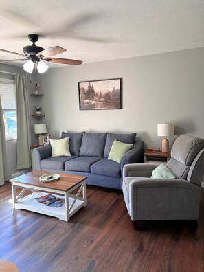 A living room with a grey sofa, a grey armchair, a coffee table, a ceiling fan, and a framed picture on the wall. - Muddy Boots HideoutI Cozy 2BR I 15m to RRG I Wi-Fi (Stanton)