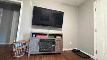 Ein Zimmer mit einem an der Wand befestigten Flachbildfernseher, einem Holzkabinett mit integriertem Kamin und einem kleinen runden Tisch mit Metallgitter.
