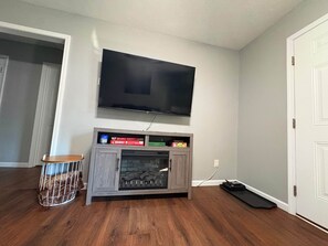 Ein Zimmer mit einem an der Wand befestigten Flachbildfernseher, einem Holzkabinett mit integriertem Kamin und einem kleinen runden Tisch mit Metallgitter.