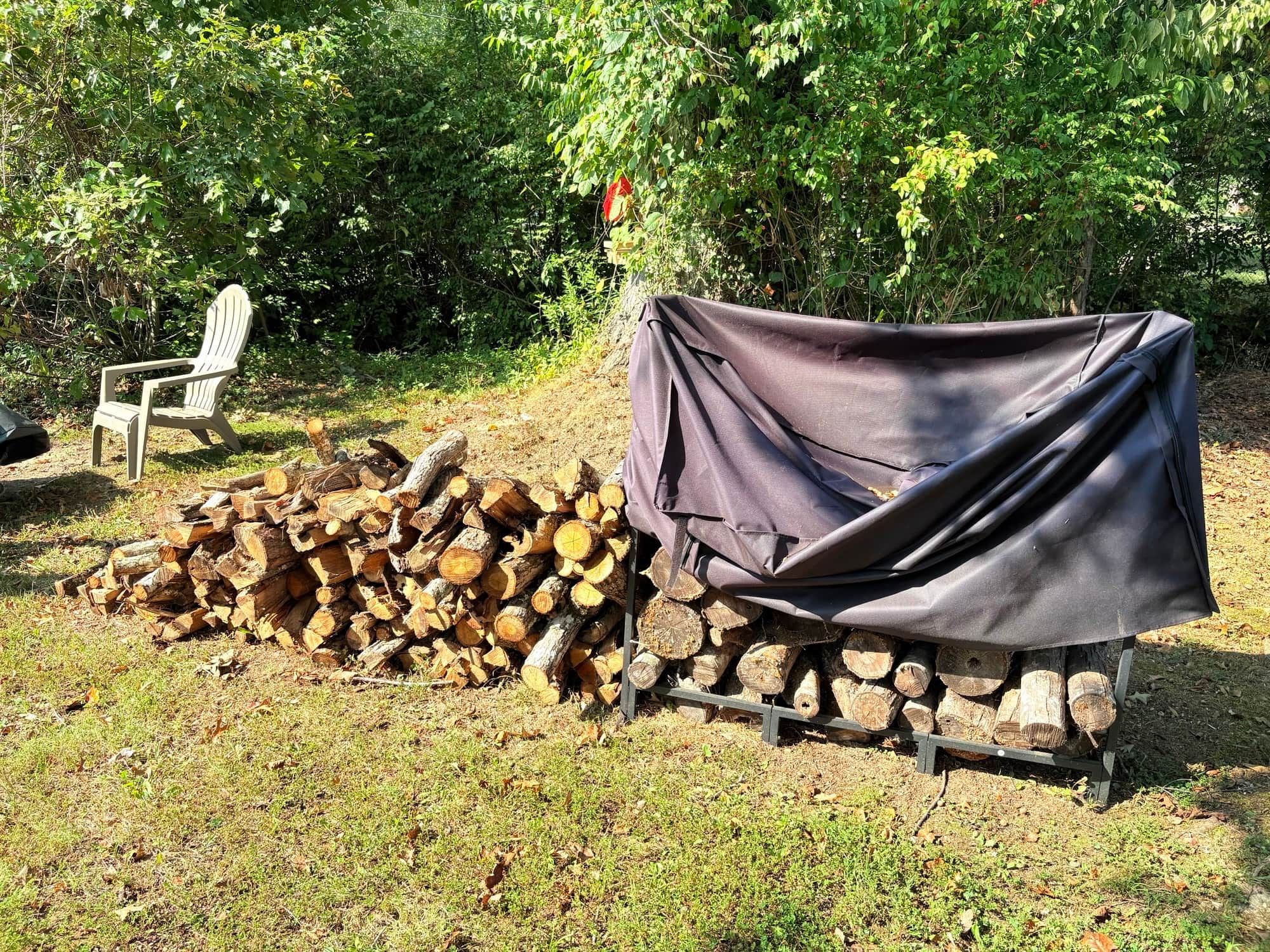 A pile of firewood covered with a tarp, a covered grill, and a chair in a grassy outdoor area.