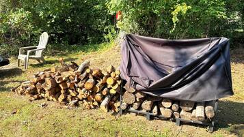 A pile of firewood covered with a tarp, a covered grill, and a chair in a grassy outdoor area.