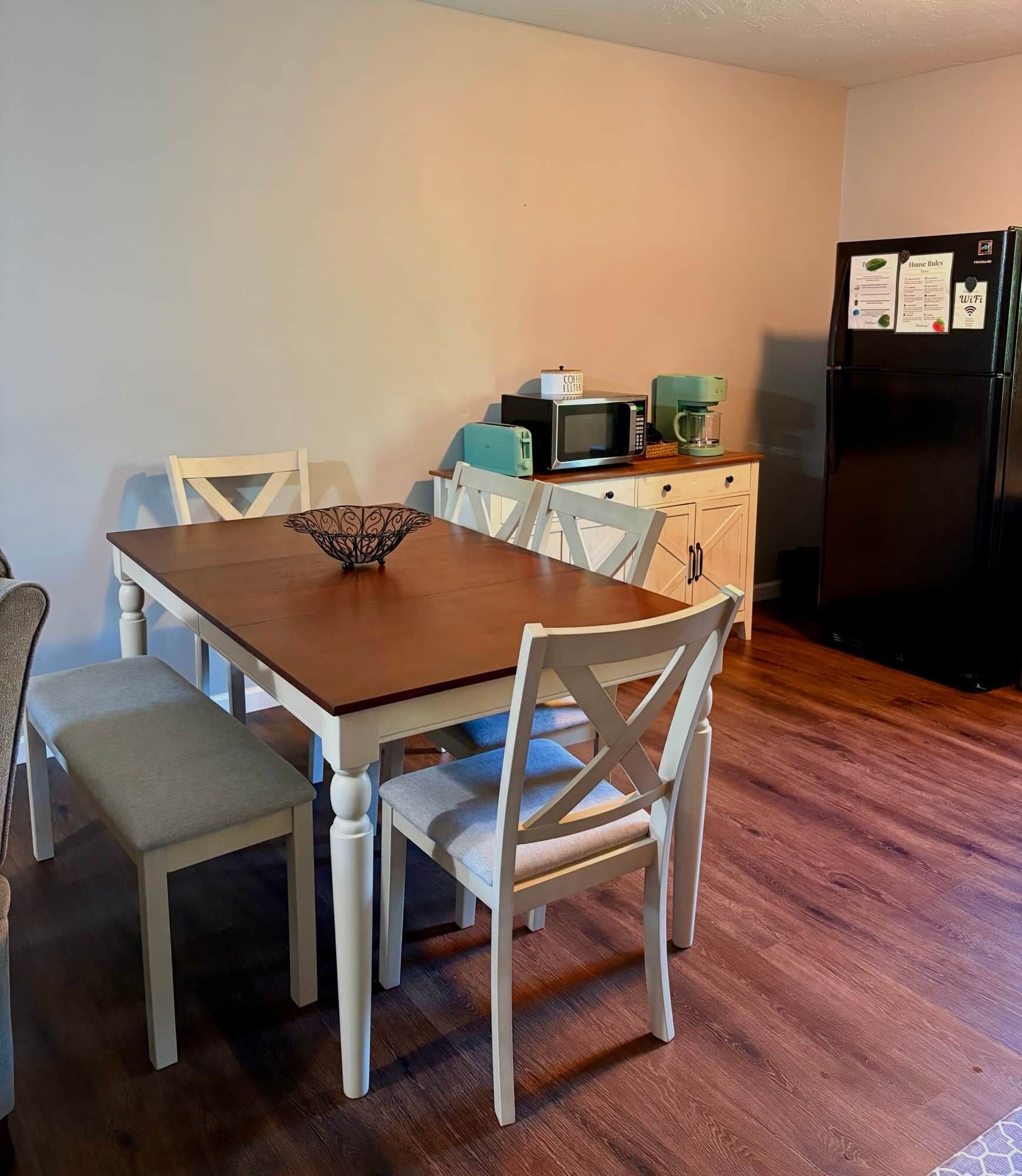 A dining area with a wooden table and white chairs, a coffee maker, and a refrigerator with a WiFi symbol.