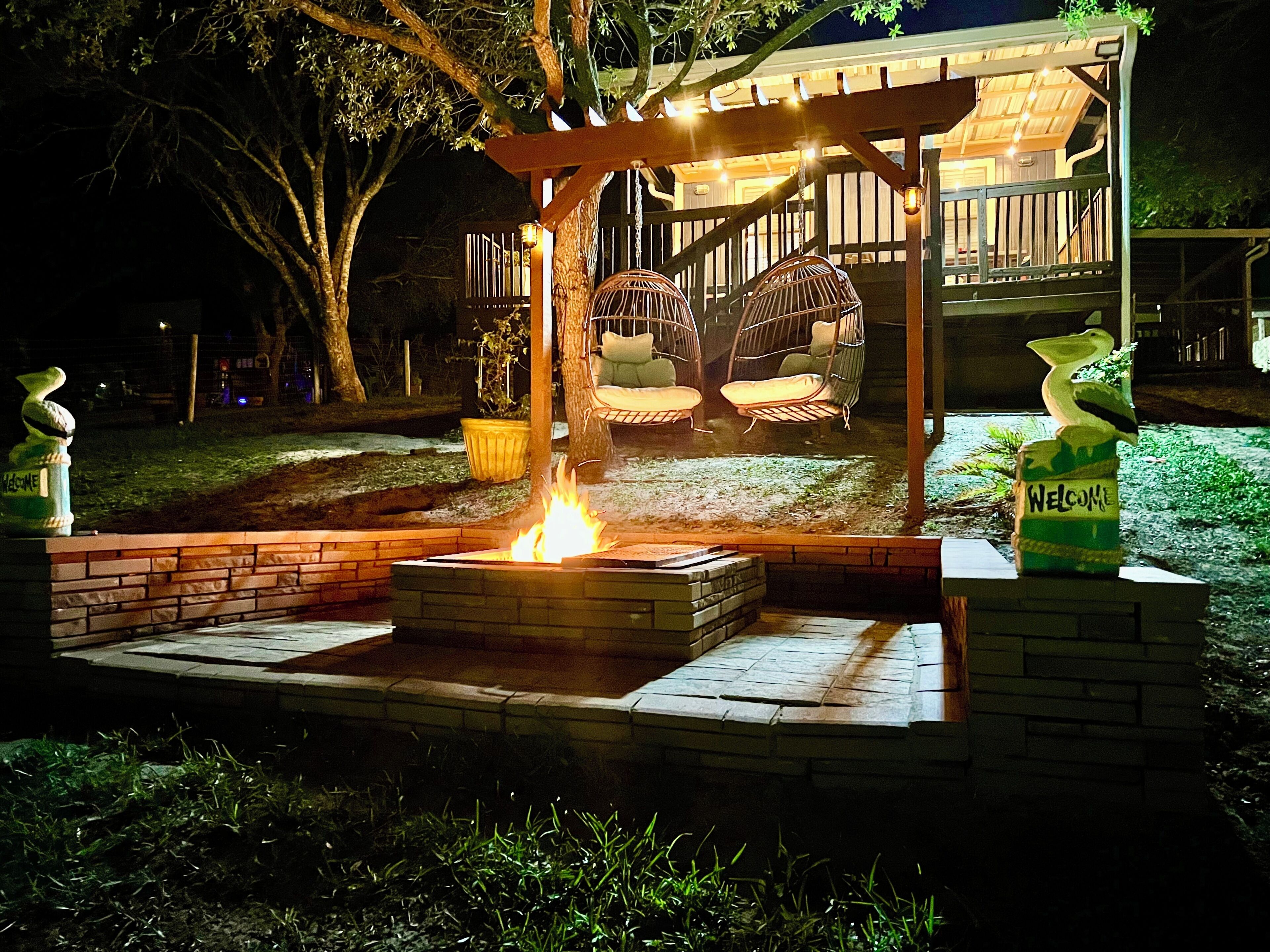 A fire pit surrounded by brick seating, a pergola with hanging chairs, and a welcoming sign.
