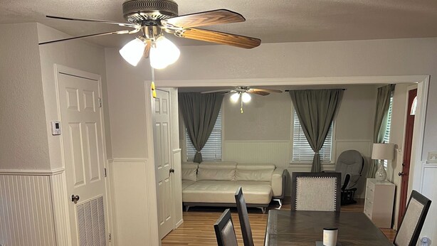 A dining area with a wooden table and chairs, a ceiling fan, and a view into a living room with a sofa and curtains.