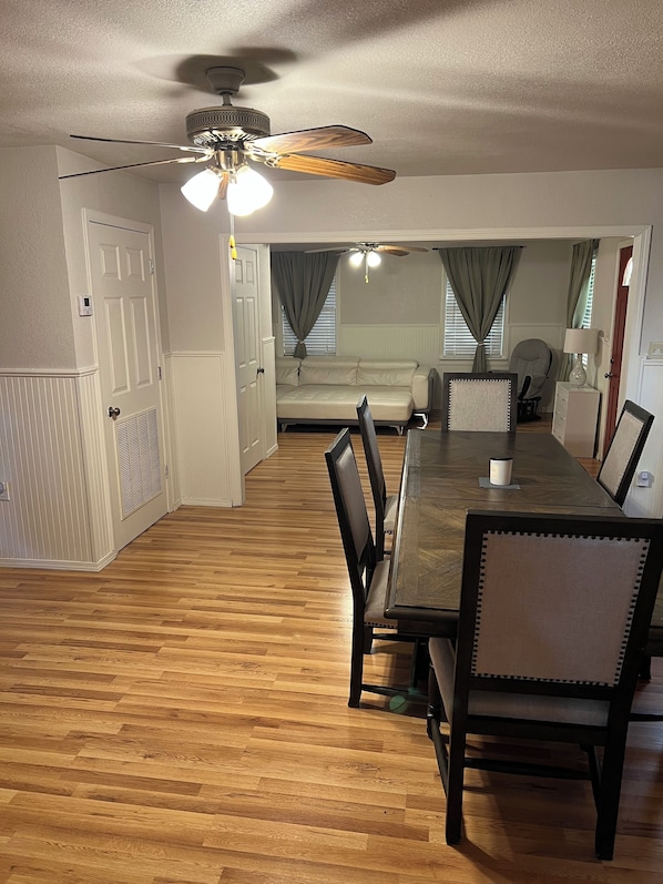 A dining area with a wooden table and chairs, a ceiling fan, and a view into a living room with a sofa and curtains.