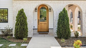 A house with a brick facade, a wooden front door, and two symmetrical evergreen trees. - Spacious New-Construction Home in Charming Bentonville! Prime location! (Bentonville)