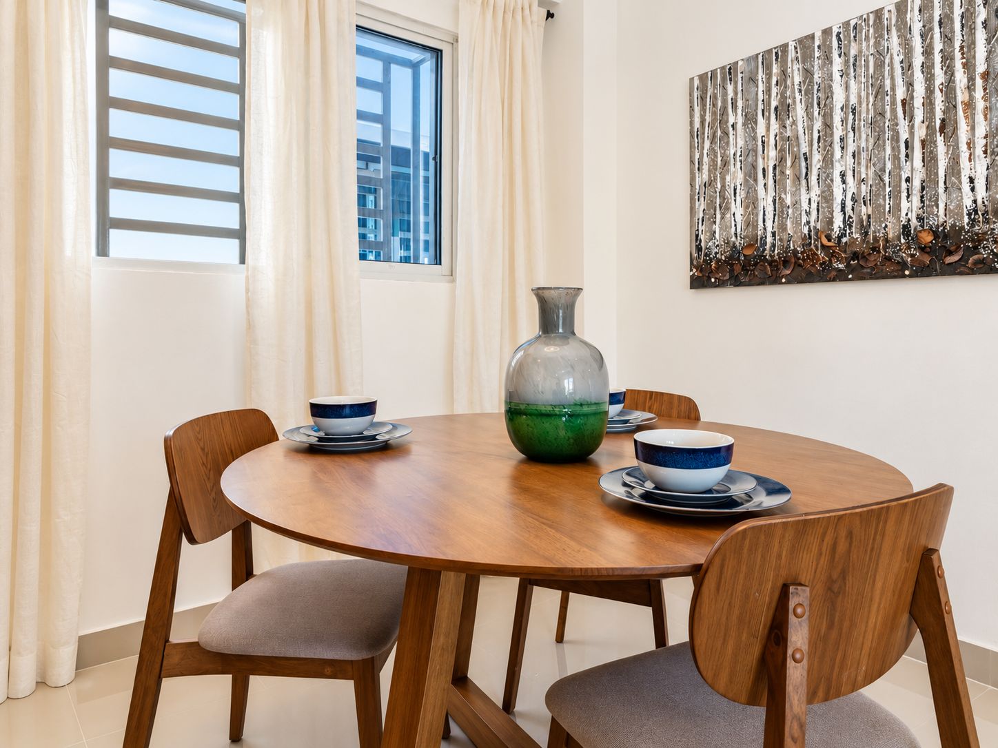 A dining area with a round wooden table, four chairs, a vase, and a painting on the wall.