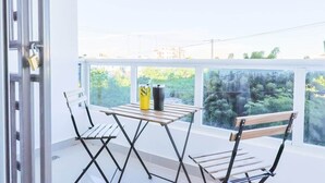 A balcony with a table and two chairs, a window with a view of greenery, and a door with a padlock.