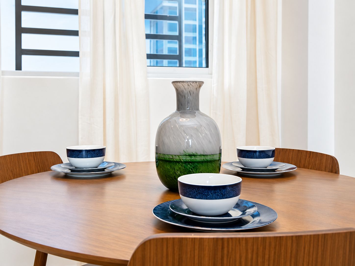 A dining table with a vase and two bowls on plates, set against a window with curtains.