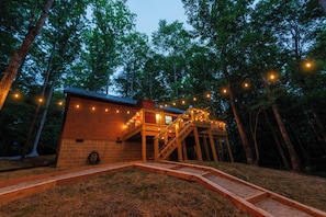 Une cabane en bois avec un escalier, entourée d’arbres et illuminée de guirlandes lumineuses.