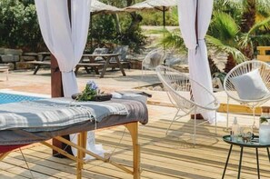 Eine Holzterrasse mit einer Massageliege, weißen Korbstühlen und einem kleinen runden Tisch mit Flaschen und einem Glas.