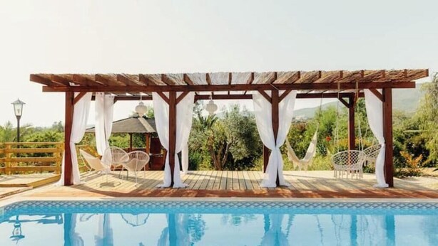 Área de piscina com pergolado de madeira, cortinas brancas e cadeiras brancas.
