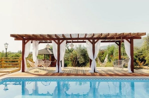Ein Poolbereich mit einer Holzpergola, weißen Vorhängen und weißen Korbstühlen.