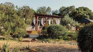 Un belvédère à la structure en bois et aux rideaux blancs, entouré d’une végétation luxuriante et d’arbres.