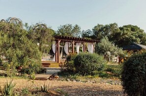 Ein Pavillon mit Holzbau und weißen Vorhängen, umgeben von dichtem Grün und Bäumen.