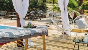 Een houten terras met een massagetafel, witte rieten stoelen en een klein rond tafeltje met flessen en een glas.