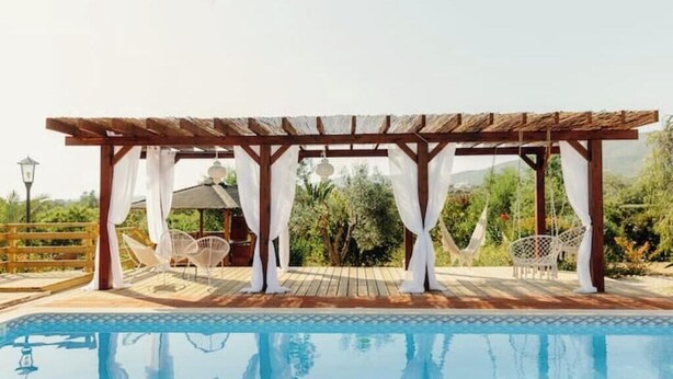 Een zwembadgedeelte met een houten pergola, witte gordijnen en witte rieten stoelen.
