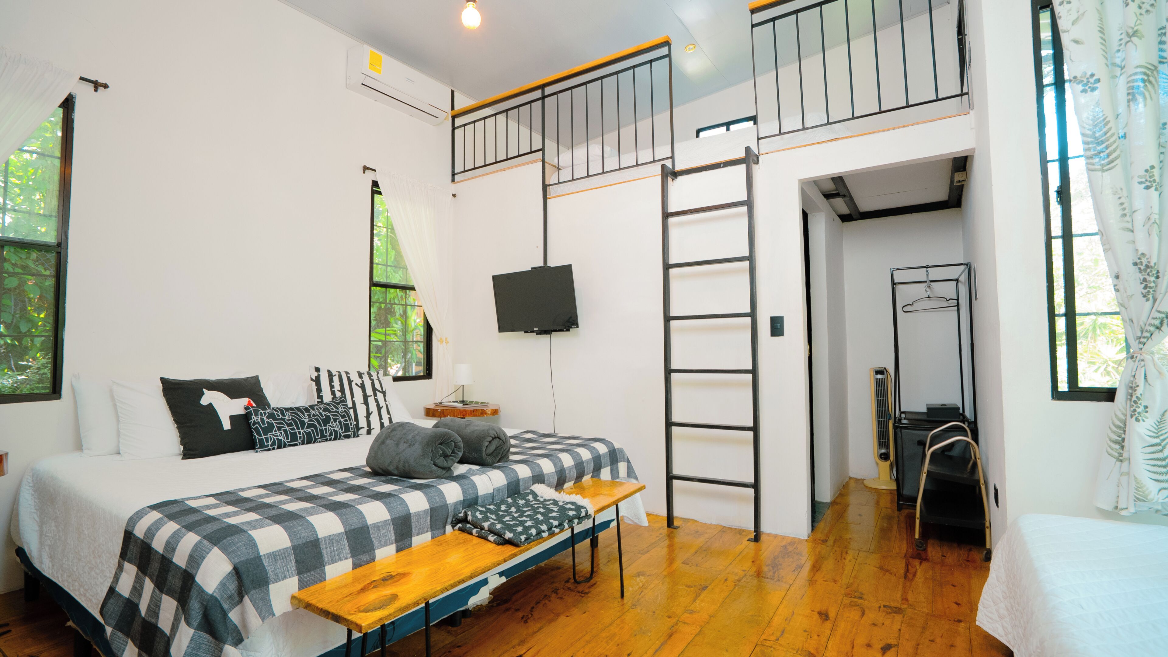 A bedroom with a bed, a desk, a TV, a ladder, and a window.