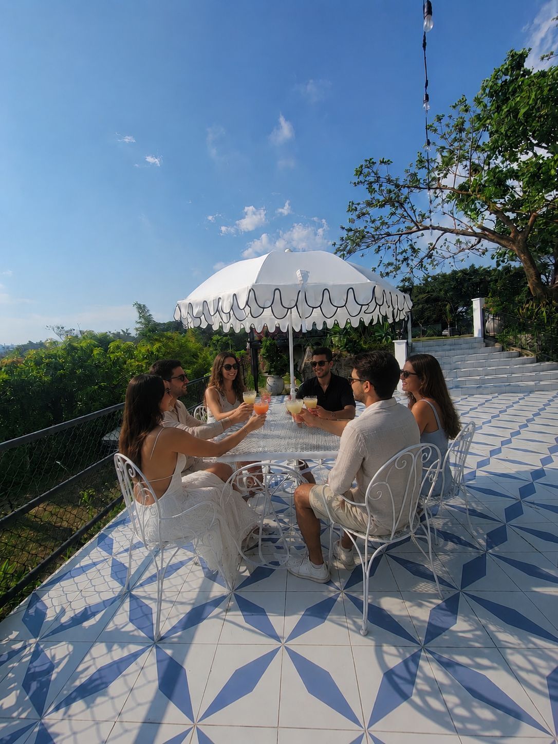 A group of people seated at a table with a white umbrella, outdoors on a tiled patio with a view of greenery and a staircase.