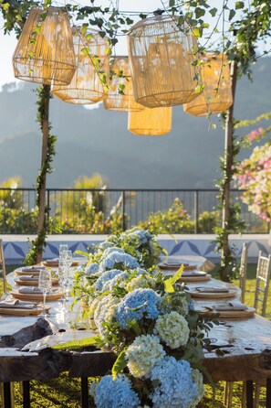 Une table dressée à l’extérieur, offrant une vue sur les montagnes, entourée de verdure et agrémentée de luminaires suspendus en osier.
