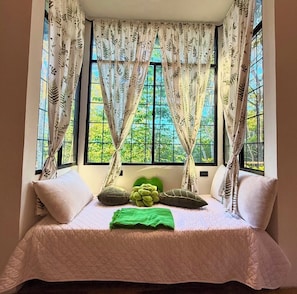 A room with a bed, a window with curtains, and a view of greenery outside.