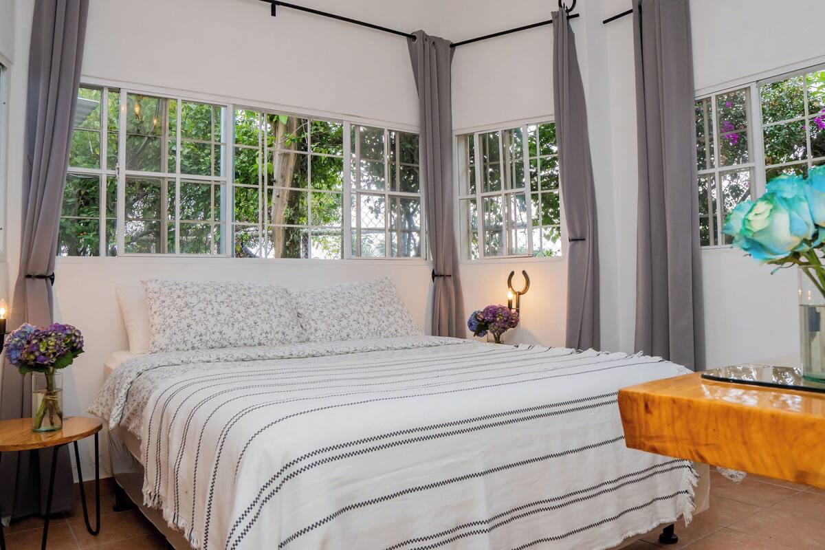 Quarto com cama, mesa de cabeceira, mesa lateral de madeira e janela com cortinas.