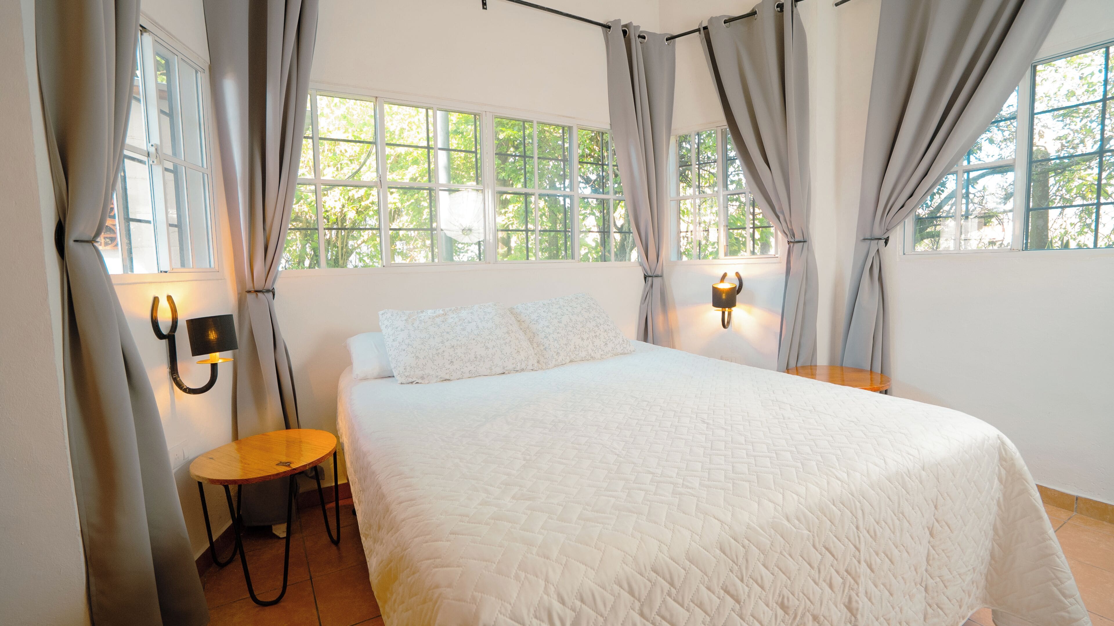 Une chambre à coucher avec un grand lit, deux tables de chevet, une lampe fixée au mur et une fenêtre avec des rideaux.